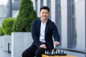 Cheerful Asian businessman playing chess in the yard smiling and looking forward to the camera photo
