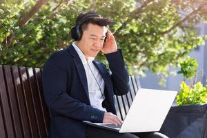 cheerful asian business man sitting on a bench in a city park in downtown and listening to music photo