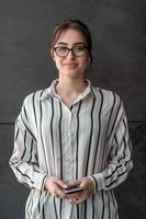 Startup businesswoman in shirt with a glasses using smartphone while standing in front of gray wall during break from work outside photo
