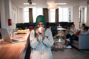 African muslim business woman wearing a green hijab drinking tea while working on laptop computer in relaxation area at modern open plan startup office. photo