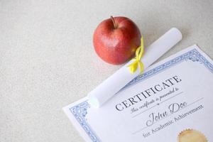 A certificate of achievement lies on table with small scroll and red apple. Education documents photo