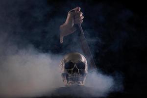 cráneo humano de halloween en una vieja mesa de madera sobre fondo negro. forma de hueso del cráneo para la cabeza de la muerte en el festival de halloween que muestra el cuchillo asesino mata la sentencia muerta, copia el espacio foto