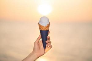 toma helado con un cono de galleta en la mano en la playa, momento del atardecer. foto