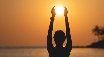 Sunset ocean view. Sun above the sea on orange sky. Woman standing on beach holding sun in her hands. photo