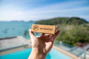 Man hand holding no smoking sign written on wood panel with beach background. photo