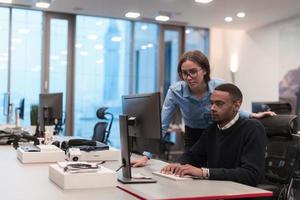 Young smiling woman explaining to serious African American coworker project strategy. Diverse startup coworkers students woman and man talking discussing working in modern office using computer. photo