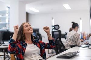 Happy casual business woman celebrating success while working on desktop computer in modern open plan startup office interior. Selective focus photo