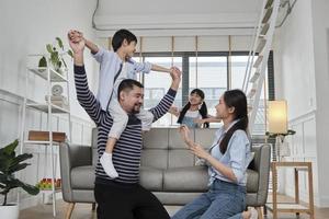 Asian Thai family together, dad plays and teases with children and mum by carrying and holding son on shoulders in home living room, happy leisure times, lovely weekend, wellbeing domestic lifestyle. photo