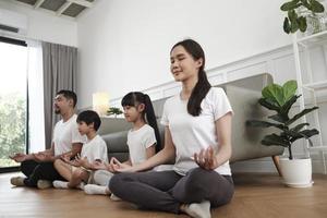 feliz familia tailandesa asiática, padres e hijos se sientan en el piso de la sala de estar, cierran los ojos juntos, meditan tranquilamente en posición de loto y practican yoga saludable, fin de semana de estilo de vida doméstico. foto