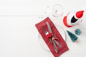 Christmas table setting with white dishware, silverware and red and green decorations on white wooden background. Top view. photo