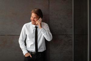 Empresario de inicio en una camisa blanca con una corbata negra usando un teléfono inteligente mientras está de pie frente a la pared gris durante el descanso del trabajo afuera foto