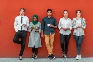 grupo multiétnico de empresarios casuales que usan un teléfono inteligente durante un descanso para tomar café del trabajo frente a la pared roja afuera. foto