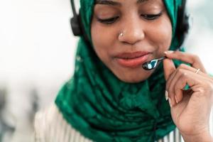 Afro Muslim female with green hijab scarf customer representative business woman with phone headset helping and supporting online with customer in modern call centre photo