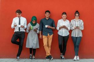 Multiethnic group of casual businesspeople using smartphone during a coffee break from work in front of the red wall outside. photo