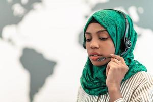 Afro Muslim female with green hijab scarf customer representative business woman with phone headset helping and supporting online with customer in modern call centre photo
