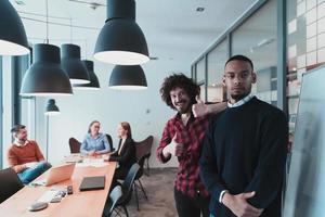 Portrait of two happy millennial male business owner in modern office. Two businessman smiling and looking at camera. Busy diverse team working in background. Leadership concept. Head shot. photo