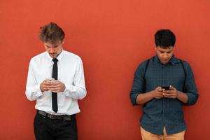 Casual multiethnic startup business men one of them is Indian using smartphone during break from work in front of red wall outside photo