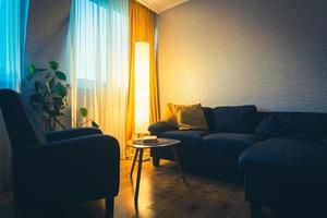 acogedor salón de apartamento con sofá y almohadas amarillas y mesa estilizada con libro de flores y ventana con luz azul exterior en invierno. lectura de libros temprano en la noche. copiar pegar pared foto