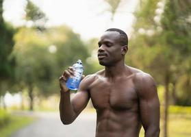 atractivo deportista africano cansado y sediento después de hacer ejercicio bebiendo agua. atletas hombre africano descansando después de entrenar corriendo en el parque del jardín. concepto de hombre deportivo. foto