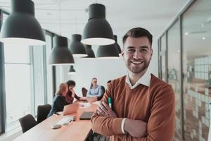 retrato de un feliz dueño de un negocio milenario en una oficina moderna. hombre de negocios con gafas, sonriendo y mirando a la cámara. equipo diverso ocupado trabajando en segundo plano. concepto de liderazgo. disparo a la cabeza. foto