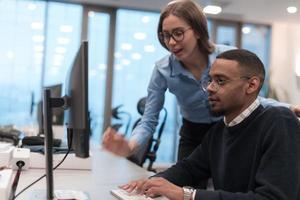 Young smiling woman explaining to serious African American coworker project strategy. Diverse startup coworkers students woman and man talking discussing working in modern office using computer. photo