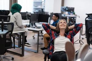 Casual business woman taking a break with legs on her table while working on desktop computer in modern open plan startup office interior. Selective focus photo