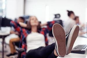 Casual business woman taking a break with legs on her table while working on desktop computer in modern open plan startup office interior. Selective focus photo