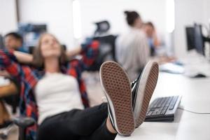 Casual business woman taking a break with legs on her table while working on desktop computer in modern open plan startup office interior. Selective focus photo