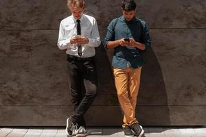 Casual multiethnic startup business men one of them is Indian using smartphone during break from work in front of grey wall outside photo