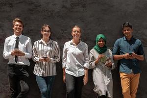 grupo multiétnico de empresarios casuales que usan teléfonos móviles durante la pausa para el café del trabajo frente a la pared negra afuera foto