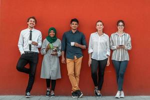 Multiethnic group of casual businesspeople using smartphone during a coffee break from work in front of the red wall outside. photo
