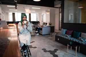 African muslim business woman wearing a green hijab drinking tea while working on laptop computer in relaxation area at modern open plan startup office. photo