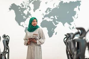 Portrait of muslim female software developer with green hijab holding tablet computer while standing at modern open plan startup office