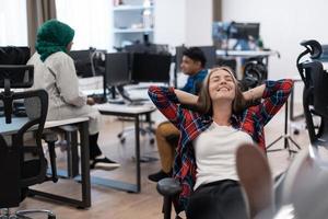 Casual business woman taking a break with legs on her table while working on desktop computer in modern open plan startup office interior. Selective focus photo