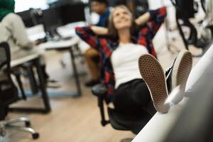 Casual business woman taking a break with legs on her table while working on desktop computer in modern open plan startup office interior. Selective focus photo