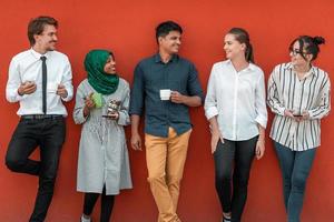 Multiethnic group of casual businesspeople using smartphone during a coffee break from work in front of the red wall outside. photo