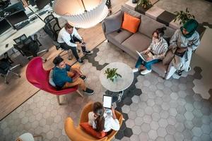 Top view photo of Multiethnic startup business team having brainstorming in relaxation area of modern office interior working on laptop and tablet computer