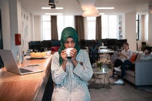 African muslim business woman wearing a green hijab drinking tea while working on laptop computer in relaxation area at modern open plan startup office. photo