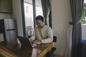 un joven asiático pasa su tiempo en casa sentado en el comedor trabajando felizmente en su computadora portátil foto