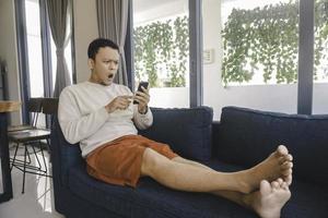 A young Asian man spends his free time at home sitting on a cozy sofa in the living room shocked by looking his smartphone photo