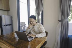 un joven asiático pasa su tiempo en casa sentado en el comedor trabajando felizmente en su computadora portátil foto