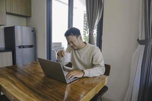 un joven asiático pasa su tiempo en casa sentado en el comedor trabajando en su computadora portátil y se siente frustrado foto