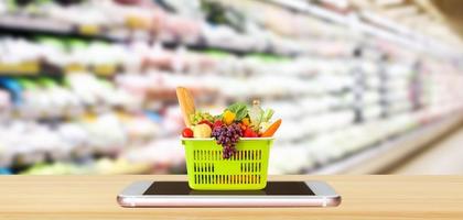 alimentos frescos y verduras en la cesta de la compra en el teléfono inteligente móvil en la mesa de madera con pasillo de supermercado fondo borroso concepto en línea de comestibles foto