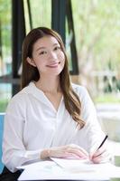 Asian confident woman wear a white shirt is using a notepad to work and there are many documents on the table. her face with smiling in a working room at home. photo