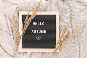 Autumnal Background. Black letter board with text phrase Hello Autumn and dried grass lying on white knitted sweater. Top view, flat lay. Thanksgiving banner. Hygge mood cold weather concept photo