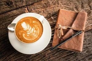 una taza de café caliente con arte latte en la superficie con el libro de cuero sobre la mesa de madera. foto