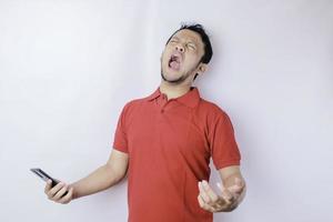 un joven asiático enojado se ve descontento usando una camisa roja con expresiones faciales irritadas sosteniendo su teléfono foto