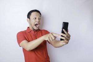 Surprised Asian man wearing red t-shirt pointing at his smartphone, isolated by white background photo