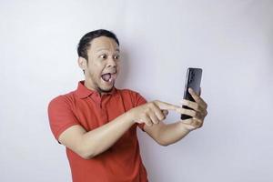 Surprised Asian man wearing red t-shirt pointing at his smartphone, isolated by white background photo