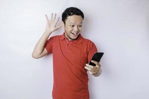 hombre asiático sorprendido con una camiseta roja apuntando a su teléfono inteligente, aislado de fondo blanco foto